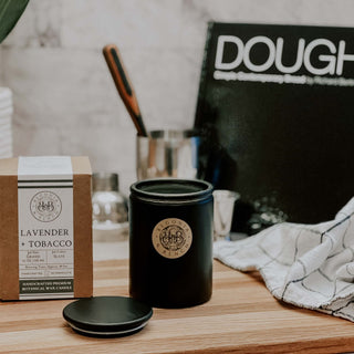 A black scented candle with a matching lid sits on a wooden surface next to its brown packaging labeled "Lavender + Tobacco™ - Signature Aroma by Waybread". Behind it, blurred kitchen utensils including a knife and a towel are visible. A cookbook titled "DOUGH" partially appears in the background. A checkered cloth is to the right.