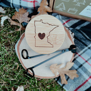 A Minnesota Engraved Bamboo Lid featuring the outline of Minnesota and a heart symbol rests on a wooden slice, accompanied by black candle wick trimmers. Nearby, a scented candle adds warmth to the setup on a plaid blanket surrounded by dried autumn leaves and grass, with a box in the background.