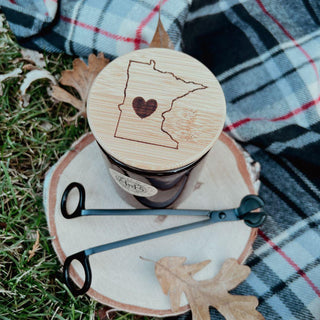 The Minnesota Engraved Bamboo Lid, etched with the outline of Minnesota and a heart, tops this scented candle that rests on a wooden slice beside a wick trimmer and snuffer. The scene is set against grass, dry leaves, and a plaid blanket, evoking an autumnal, cozy atmosphere.