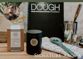 Image of the Lavender + Tobacco™ - Signature Aroma by Waybread candle and its packaging, surrounded by various kitchen utensils. The candle label reads "Lavender + Tobacco." In the background, a "DOUGH" branded box is visible. The text "Signature Aroma" is at the bottom. A circular inset shows a man's face with sunglasses and the word "Waybread." This fall candle promises a cozy, aromatic experience.