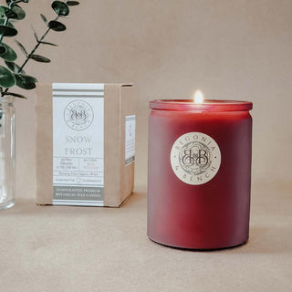 A lit "Snow Frost™" candle from Begonia & Bench® in a glass jar sits on a beige surface. Behind it, a cardboard box reveals this 12oz artisan candle's blend of Grand Juniper & Red Velvet. A small vase with green leaves is partially visible on the left.