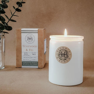 A lit Teakwood & Fig™ 12oz Finely Crafted Scented Candle by Begonia & Bench®, featuring a minimalist white label, is placed next to its brown box packaging that also highlights essential oils and a cotton wick. In the background, a small clear jar holds green foliage against a plain surface.