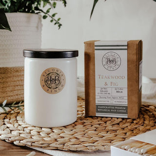 Image showcasing the Teakwood & Fig™ 12oz Finely Crafted Scented Candle by Begonia & Bench® in a white jar with a black lid, next to its brown packaging labeled "Begonia & Bench." The label on the packaging details "Teakwood & Fig," "Jet Set," "Grand," and "Weight: Approximately 12 oz / 345 ml." Both items are placed on a woven placemat.