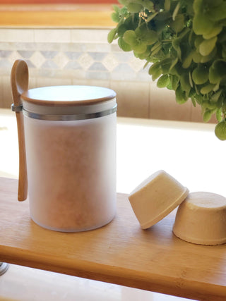 A frosted glass jar topped with a Bamboo Lid, reminiscent of a fall candle, rests on a light wooden surface. Adjacent to it are two white, dome-shaped tablets. In the background, a leafy green plant cascades down, with a tiled wall partially in view. The setting suggests it could be either a bathroom or kitchen.