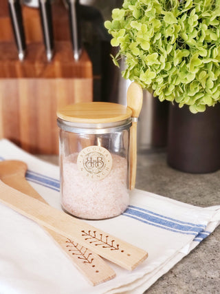A glass jar labeled "Begonia Botanica" filled with pink Himalayan salt sits on a countertop. It has a wooden lid and an attached Bamboo Spoon & Clip. Next to it are wooden utensils with floral engravings resting on a white cloth with blue stripes. A green potted plant and a fall candle are in the background.