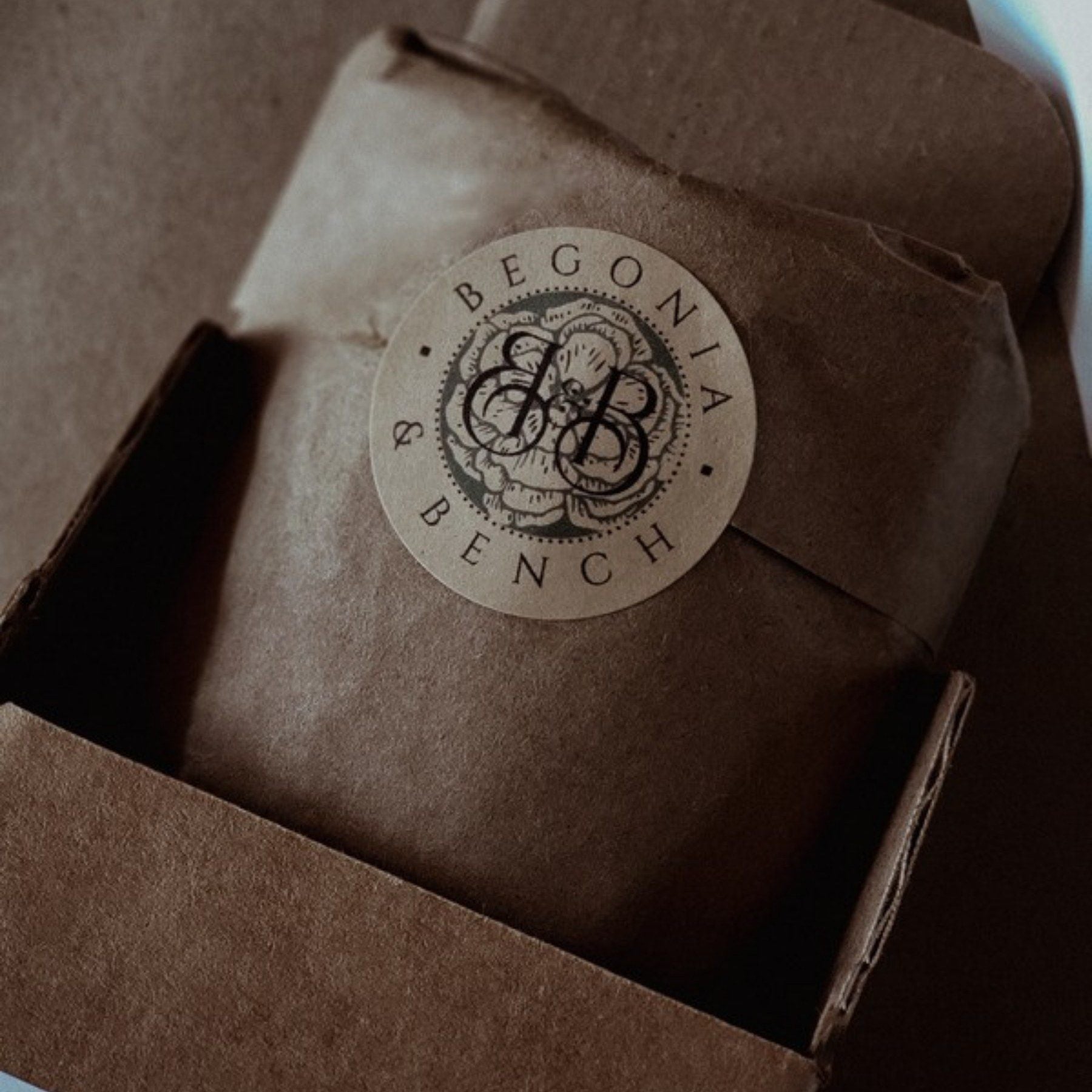 A candle wrapped in kraft paper inside an open kraft box, closed with the round Begonia and Bench seal