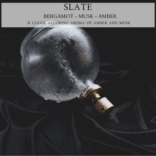A frosted glass perfume bottle with a partially visible gold cap lies on a black, velvety cloth. The top section of the image contains text that reads: "Slate™ - 9oz. Amber Jar Candle - By Begonia & Bench®. Bergamot - Musk - Amber. A clean, alluring citrus amber musk blend." The dark setting highlights the elegance of the bottle.