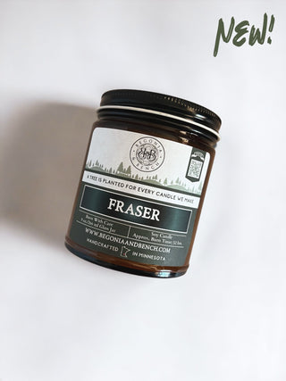 A Begonia & Bench® Fraser™ 9oz. amber jar candle, featuring a scenic white label with pine trees and the text "A tree is planted for every candle we make." The label also includes burn time and the phrase "handcrafted in Minnesota." A handwritten "NEW!" note in green decorates the top right corner.