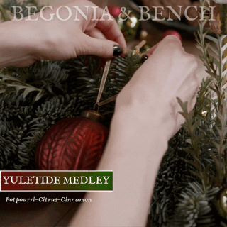 Close-up of someone tying a red bauble ornament onto a Christmas tree branch. The person's hands are adjusting the hanging ornament. Overlaid text reads, "BEGONIA & BENCH®" at the top, "YULETIDE MEDLEY™ - 16oz Twin Wick" in white on a red background, and "Festive Fragrances - Citrus - Cinnamon" at the bottom.