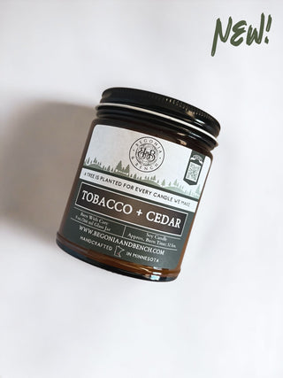 Introducing the new fall candle handcrafted in Minnesota: the Tobacco & Cedar - 9oz. Amber Jar Candle by Begonia & Bench®. This brown jar scented candle features a black lid and a label with a logo, the phrase "A tree is planted for every candle we make," and boasts a burn time of over 50 hours. Visit www.BegoniaAndBench.com for more information. "NEW!" is highlighted in green at the top right corner of the label.