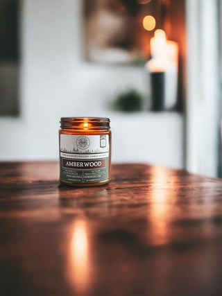 A lit Amberwood - 9oz. Amber Jar Candle by Begonia & Bench®, complete with a wooden lid, sits on a polished wooden surface. The background is softly blurred, showcasing another larger, out-of-focus candle and some decor, creating a cozy and warm ambiance perfect for fall.