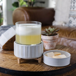 A Classic 2-in-1 Candle Warmer with a yellow and white gradient candle rests in a ribbed ceramic holder on wooden legs atop a round wooden table. In proximity, there's a small dish containing soap bars alongside a potted succulent. A brown leather couch and decorative textiles are visible in the softly focused background.