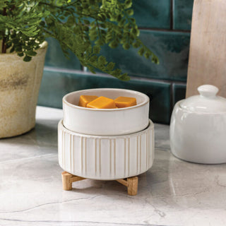 A Classic 2-in-1 Candle Warmer with a ribbed white ceramic design sits on a wooden stand, holding fragrant orange wax melts. Placed on a marble countertop with a green tile backsplash, it complements the nearby potted plant and elegant white jar candle.