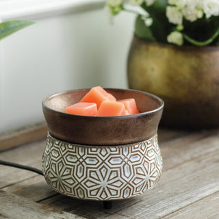 A *Classic 2-in-1 Candle Warmer* featuring a geometric pattern sits on a wooden surface, filled with fragrant, vibrant orange wax melts. In the background, a brass-colored pot holds white flowers, while a green plant peeks from the left. The candle warmer is plugged in to provide instant ambiance.