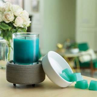 A turquoise jar candle sits elegantly on the Classic 2-in-1 Candle Warmer, its charm illuminated by a round, textured holder. Beside it, a white bowl overflows with wax melts in harmonious shades, with a few artfully scattered across the wooden surface. In the softly blurred background, white roses in a vase and green chairs enhance the serene ambiance of the bright room.