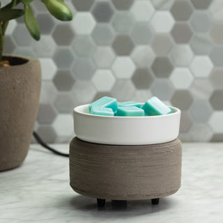 A hexagonal tile backsplash highlights a Classic 2-in-1 Candle Warmer, characterized by its textured base and smooth white dish resting on a gray marble countertop. The dish holds fragrant blue wax melts. To the left, part of a potted plant adds a natural touch to the scene.
