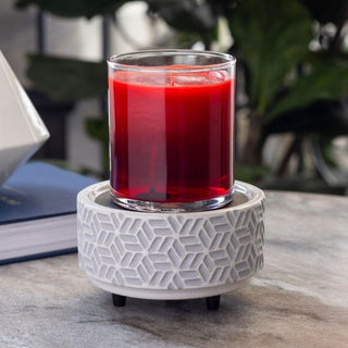 A red candle in a clear glass jar is placed on the Classic 2-in-1 Candle Warmer, which features a geometric pattern. This setup rests on a marble surface accompanied by a blue book. In the softly-focused background, green plants contribute to creating a cozy, natural atmosphere filled with warmth and charm.