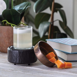 A glass of white liquid rests on a black wooden coaster. Nearby, caramel candies spill out from a tilted brown bowl, their sweet aroma blending with the scent from the Classic 2-in-1 Candle Warmer. In the background, two stacked books accompany a green plant on a light wooden surface.