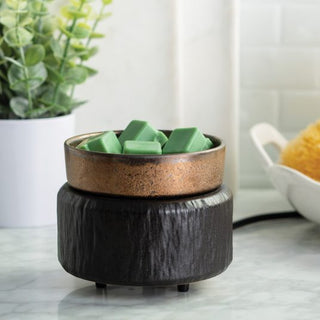 A Classic 2-in-1 Candle Warmer in dark brown is placed on a marble countertop, its rustic bronze-colored bowl melting fragrance wax melts. In the background, you can see a small potted plant with green foliage alongside a white ceramic dish, suggesting the setting is either a kitchen or bathroom.