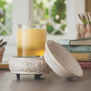 A lit yellow candle is placed in a decorative glass holder on a Classic 2-in-1 Candle Warmer featuring an ornate floral design. The warmer's lid is set beside it, accompanied by an assortment of fragrance wax melts. In the background, blurred greenery and books on a wooden table create a cozy atmosphere.