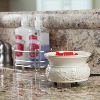 A white Classic 2-in-1 Candle Warmer adorns a granite countertop, softly dispersing the aroma of fragrance wax melts. It is plugged in and contains red wax cubes. In the background, two bottles of hand soap are placed in a metal holder next to a silver faucet.