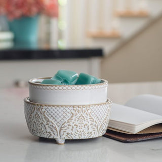 A decorative ceramic bowl with intricate patterns holds several green coffee pods. Next to the bowl, a Classic 2-in-1 Candle Warmer gently melts fragrance wax, filling the air with a soft aroma. The scene is set on a white countertop beside an open notebook, against blurred decor and a staircase backdrop.