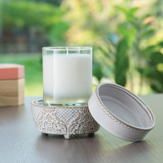 The Classic 2-in-1 Candle Warmer, featuring a white candle in a clear glass holder, rests on a decorative ceramic base with a matching lid placed beside it. The scene is complemented by blurred greenery and a wooden box with a peach-colored lid for storing fragrance wax, all elegantly arranged on top of a wooden table.