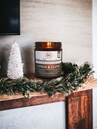 A lit 'Citrus & Clove' scented candle in an amber jar sits on a wooden mantle adorned with green pine garland and a small white Christmas tree decoration. The candle label features a holiday-themed design and text about the company's tree-planting initiative and candle details.