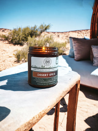 A lit Desert Spice™ - 9oz. Amber Jar Candle by Begonia & Bench® is placed on a wooden table outdoors. The background features a desert landscape with sparse vegetation, a blue sky, and a cushioned seating area with pillows under a wooden shade structure. The candle jar includes text and branding details.