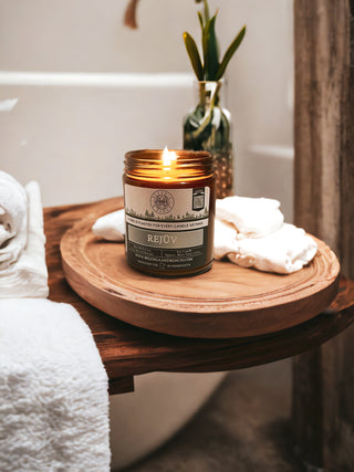 A lit Rejüv™ 9oz. Amber Jar Candle by Begonia & Bench® sits on a round wooden tray beside folded white towels. Behind the tray is a white bathtub and a vase with green foliage, creating a tranquil and spa-like ambiance.