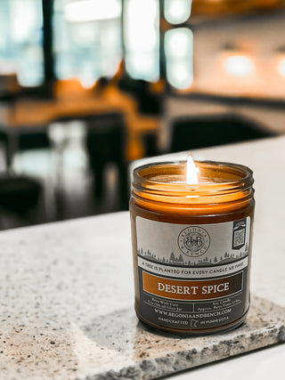 A lit Desert Spice™ 9oz. Amber Jar Candle by Begonia & Bench® sits on a speckled white countertop. The brown glass jar features a label that reads "DESERT SPICE," along with "With Cardamom," "Handcrafted," and "A tree is planted for every candle we make." In the background, a blurred indoor area with furniture creates a cozy ambiance.