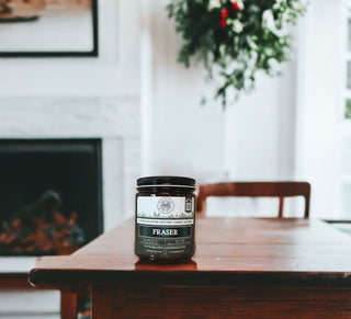 A black jar labeled "Fraser™ - 9oz. Amber Jar Candle - By Begonia & Bench®" sits on a wooden table in a cozy, light-filled room. The background features a fireplace with white trim and a lush, green plant hanging on the wall. The scented candle's label includes text and logos, and the overall ambiance suggests a warm, inviting setting.