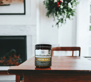A 9oz. amber jar candle labeled "Winter Sun™ - By Begonia & Bench®" is placed on a wooden table in a cozy room. The jar features a dark lid and an olive-green label, with a description elegantly positioned below the product name. In the background, a fireplace and a green plant enhance the warm, inviting atmosphere, making it the perfect setting to enjoy this delightful scented candle.