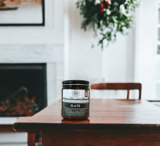 A jar of Slate™ - 9oz. Amber Jar Candle - By Begonia & Bench® sits on a wooden table. In the background, there's a white fireplace and a lush green plant hanging on the wall. The room has a bright and airy atmosphere, with sunlight streaming in through a nearby window.