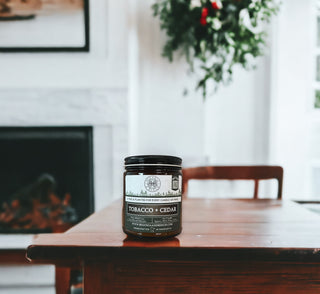 A scented candle, labeled "Tobacco & Cedar - 9oz. Amber Jar Candle - By Begonia & Bench®," sits on a wooden table in a cozy room. The label also says "Hand Poured in Charleston, SC." Behind the candle is a wooden chair, a fireplace, and greenery, creating a warm, inviting atmosphere.
