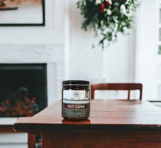 A Hot Cider™ - 9oz. Amber Jar Candle by Begonia & Bench® sits on a wooden table in a cozy setting. The fireplace in the background is not lit, and the mantel displays framed art. An ornamental plant hangs to the right, and a fall candle adds to the ambiance as soft natural light filters in through a nearby window, creating a warm and inviting atmosphere.