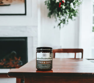 A jar labeled "Citrus & Clove - 9oz. Amber Jar Candle - By Begonia & Bench®" sits centered on a wooden table. The scene, featuring this fall candle, shows a blurred interior with a white fireplace, a framed picture, and a hanging plant with green leaves and red berries. The scene conveys a cozy, homey atmosphere.