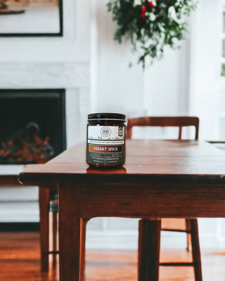 A jar of "Desert Spice™ - 9oz. Amber Jar Candle - By Begonia & Bench®" sits on a wooden dining table in a spacious, warmly lit room. The background features a white fireplace mantel adorned with a fall candle, a framed painting, and some greenery hanging. The room exudes a cozy, inviting atmosphere, with natural light filtering through windows on the right.