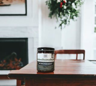 A 9oz. amber jar candle labeled "Wonderment™" by Begonia & Bench® sits on a wooden table. The background features a white fireplace with a mantel and greenery hanging on the wall, creating a cozy and inviting atmosphere. The scene is softly blurred, drawing attention to the scented candle in the foreground.