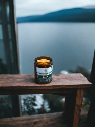 A lit P.N.W. - 9oz. Amber Jar Candle by Begonia & Bench® sits on a wooden ledge, encased in a brown jar with a green and white label reading "P.N.W." Through the window glass, the blurred view of a lake and distant mountains under a cloudy sky creates a calm, cozy, and serene atmosphere.