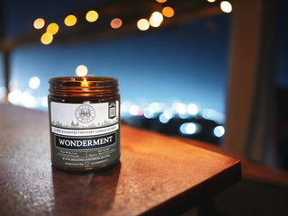 A small flame flickers atop the Wonderment™ 9oz. Amber Jar Candle by Begonia & Bench®, set on a wooden table with a blurred cityscape and string lights in the background. The packaging proudly highlights that it is handcrafted, and for each candle sold, a tree is planted, creating an inviting fall atmosphere.