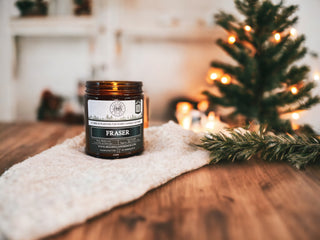 On a cozy white knit cloth, placed on a wooden table, sits a Fraser™ 9oz. Amber Jar Candle by Begonia & Bench®. In the background, there is a small Christmas tree with glowing lights and blurred candles adding to the warm ambiance, creating a festive and cozy atmosphere.