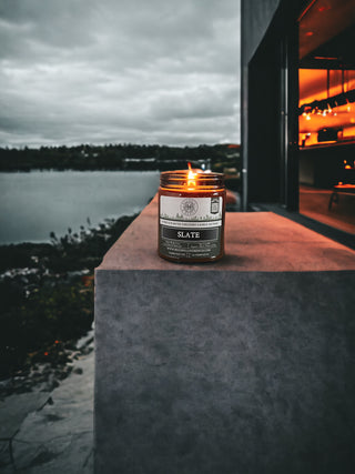 A lit Slate™ - 9oz. Amber Jar Candle - By Begonia & Bench® rests on a stone ledge, overlooking a tranquil lake bordered by dense, shadowy vegetation. To the right, a modern building with an open doorway casts a warm, orange glow. The exotic woody fragrance of the candle intermingles harmoniously with the overcast sky, striking a balance between the cool natural light and indoor warmth.