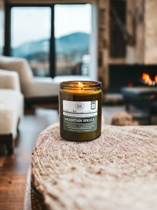 A lit "Mountain Spruce™ - 16oz Twin Wick - By Begonia & Bench®" fall candle sits on a wooden coffee table. The scented candle is in a green glass jar, displaying its name and details on the label. In the background, there is a cozy living space featuring a fireplace, an armchair, and large windows with mountain views.