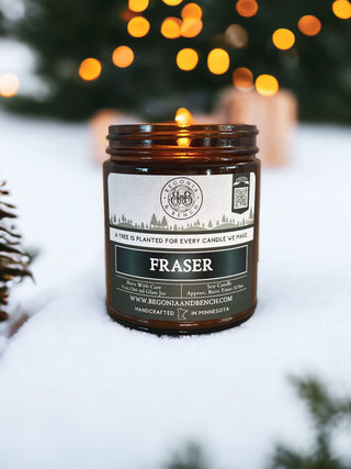 A dark green candle jar labeled "Fraser™ - 9oz. Amber Jar Candle" sits in the snow, surrounded by blurred Christmas lights in the background. The label on this scented candle indicates that a tree is planted for every candle purchased, provides burning instructions, and states that it is handcrafted in Minnesota by Begonia & Bench®.