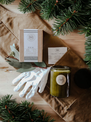 A flat lay image features a brown apron, a pair of white gloves, a green candle, and a boxed P.N.W. scented candle. Evergreen sprigs frame the composition, and a business card from "Her Own Botanical" is placed near the top right. The background is a wooden surface.