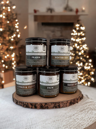 A festive display featuring five scented candles, including the "Fraser™ - 9oz. Amber Jar Candle - By Begonia & Bench®," set on a wooden slab. The candles are labeled "Fraser," "Winter Sun," "Citrus & Clove," "P.N.W.," and "Smoky Pine." The warmly lit background showcases a decorated Christmas tree with a blurred mantle adorned with holiday decor, creating a cozy holiday ambiance.