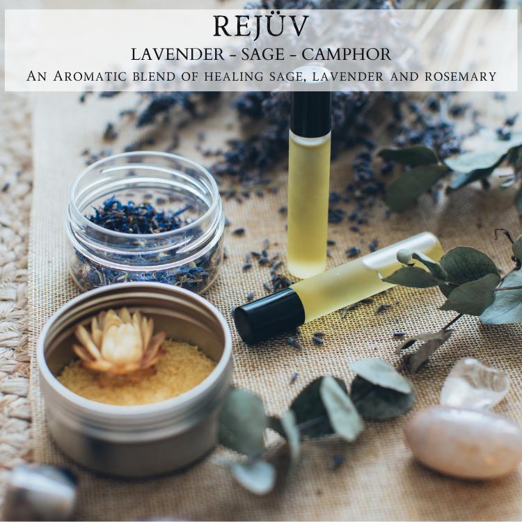 A serene display of wellness products features a glass jar with dried lavender, an open tin of powder adorned with a wooden flower, two roller bottles containing yellowish oil, and scattered eucalyptus leaves on a textured fabric surface. The scene is warmly lit by the "Rejüv™ - 9oz. Amber Jar Candle - By Begonia & Bench®." Text reads: "REJÜV Lavender - Sage - Camphor, An Aromatic blend of healing sage, lavender, and rosemary.