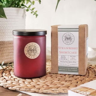 A red artisan candle in a glass jar with a black lid sits next to a brown paper bag labeled "Strawberry Shortcake - By Begonia & Bench®." Both items rest on a woven placemat. Surrounded by plants and natural wood elements, the scene evokes a cozy, rustic atmosphere.