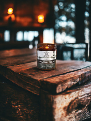 A lit candle from Begonia & Bench® labeled "Tobacco & Cedar - 9oz. Amber Jar Candle" sits on a rustic wooden table in a dimly lit room. The background is blurred but reveals warm ambient lighting, with hints of other wooden furniture and softly glowing candles, creating a cozy and inviting atmosphere.